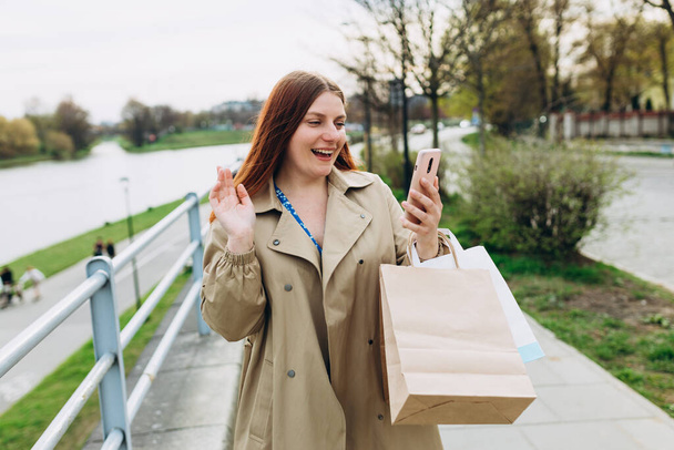 Rotschopf-Frau macht Selfie und hält Papiertüten auf Flusshintergrund, Frühlingszeit. Urban Lifestylekonzept. Fröhlich-fröhliche junge Frau läuft auf Stadtstraße. - Foto, Bild