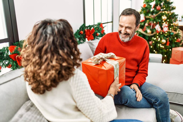 Middle age hispanic couple holding christmas gift sitting on the sofa at home. - Photo, image