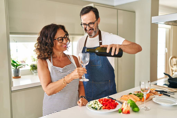 Middle age hispanic couple cooking and pouring wine on glass at kitchen - Fotó, kép