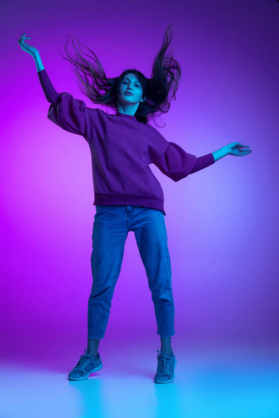 Full-length portrait of young excited girl, student jumping isolated over purple background in neon. Concept of emotions, facial expression, art, beauty - Фото, зображення