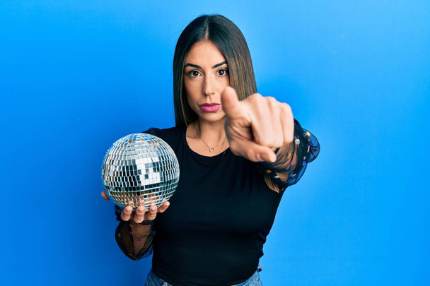 Young hispanic woman holding shiny disco ball pointing with finger to the camera and to you, confident gesture looking serious  - Fotografie, Obrázek