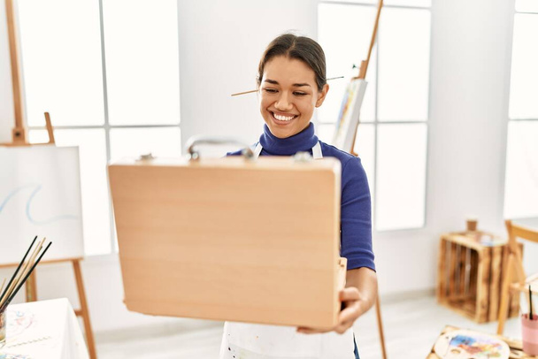 Young latin woman smiling confident opening draw briefcase at art studio - 写真・画像