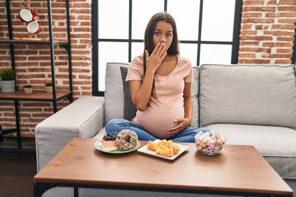 Mujer embarazada joven comiendo dulces en casa cubriendo la boca con la mano, conmocionada y temerosa de equivocarse. expresión sorprendida  - Foto, Imagen