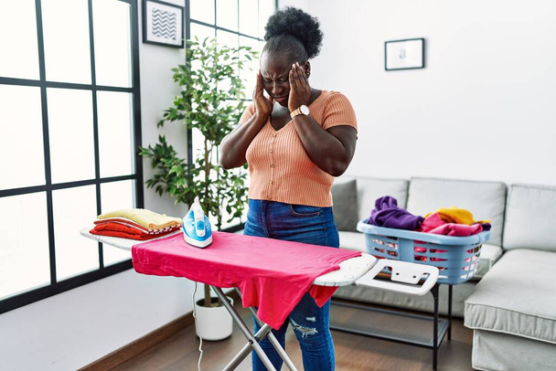 Young african woman ironing clothes at home with hand on head for pain in head because stress. suffering migraine.  - Fotó, kép