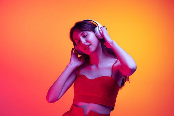 Portrait of young girl in headphones listening to music isolated over red-yellow background in neon light. Concept of emotions, facial expression, art, beauty - Fotó, kép
