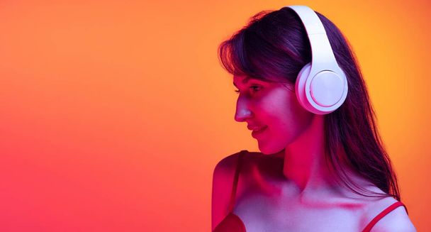 Portrait of young girl in headphones listening to music isolated over red-yellow background in neon light. Concept of emotions, facial expression, art, beauty - Фото, зображення