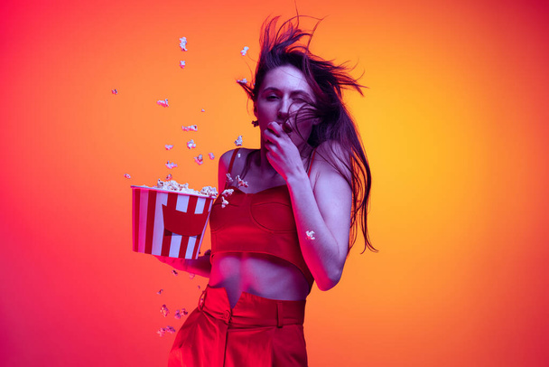 One excited young girl, student wearing 3d glasses and holding bucket of popcorn isolated on orange background in neon light, filter. - Foto, Imagen