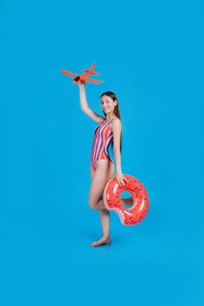 Portrait of attractive brunette woman in colored swimsuit holding rubber rings, and parper plane posing sexy on camera. Summer trendy beach style, vacation at the resort. Studio on a blue background - Photo, Image