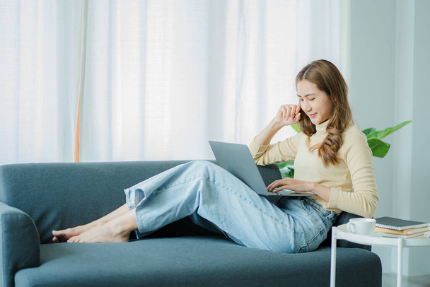 Ασιάτισσα που κάθεται στον καναπέ και εργάζεται online με την εργασία laptop στο σπίτι έννοια - Φωτογραφία, εικόνα