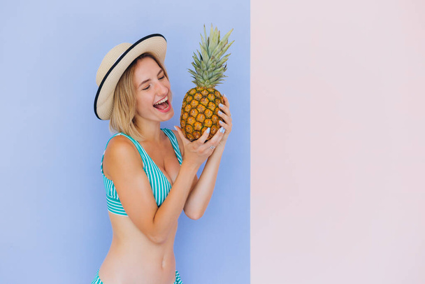 Vrolijk jong meisje op het strand met een ananas. Het meisje is gekleed in een gekleurd zwempak. Lifestyle vakantie concept van zomervakantie mensen. Houdt vers ananasfruit. Foto op een roze en blauwe achtergrond - Foto, afbeelding