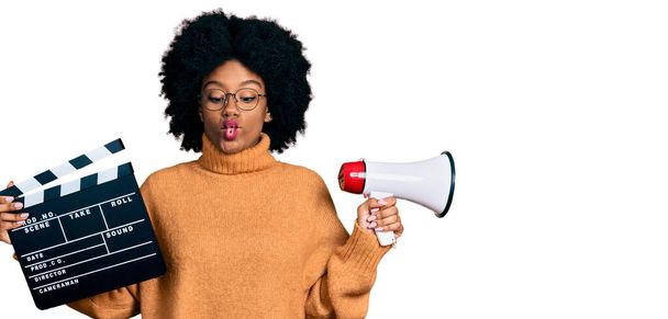 Young african american woman holding video film clapboard and megaphone making fish face with mouth and squinting eyes, crazy and comical.  - Photo, Image
