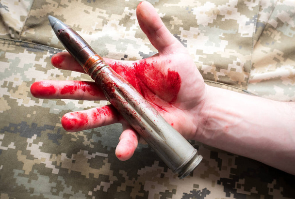 カーキ色のジャケットを背景に人間の手に血を持つシェルミサイル。戦争も軍隊の概念も. - 写真・画像