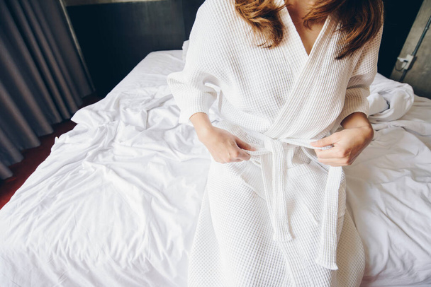Cropped shot view of young woman sitting on the bed and tying up belt of white bathrobe before taking a bath or spa massage. - Φωτογραφία, εικόνα