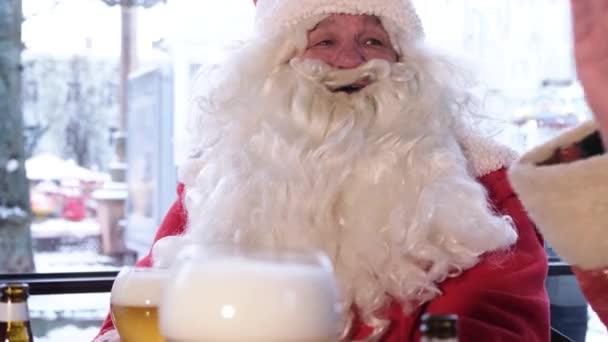Санта-Клаус с большой белой бородой разговаривает с друзьями в кафе, держа стакан пива.  - Кадры, видео