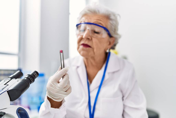 Senior grey-haired woman wearing scientist uniform holding pill at laboratory - Foto, afbeelding