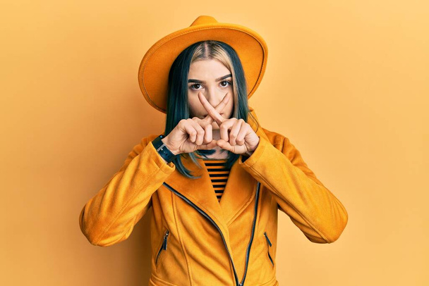 Joven chica moderna con sombrero amarillo y la expresión de rechazo chaqueta de cuero cruzando los dedos haciendo signo negativo  - Foto, Imagen