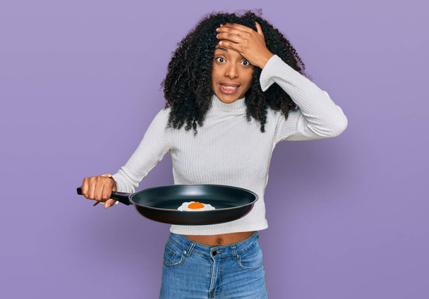 Genç Afro-Amerikalı kız elinde tavayla yumurta tutarken gergin ve sinirli, şaşırmış ve sinirli bir yüz ifadesi vardı.  - Fotoğraf, Görsel