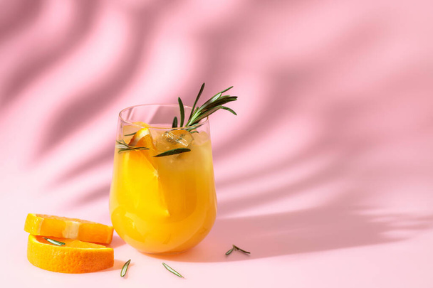 Summer cocktail with orange, rosemary, and ice. Drink on pink background with palm leaf shadow. Summer, tropical, fresh cocktail concept. - Foto, imagen