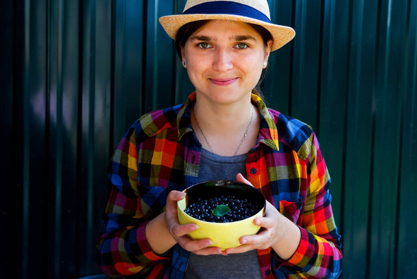Defocus young woman in a hat and shirt smiles and shows a basket of blueberry and bilberry. Dark green steel background. Summer food, fruits. Summer background. Out of focus. - Photo, Image