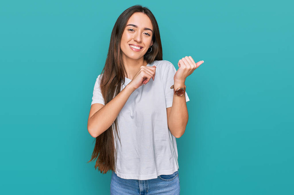 Young hispanic girl wearing casual white t shirt pointing to the back behind with hand and thumbs up, smiling confident  - Fotó, kép