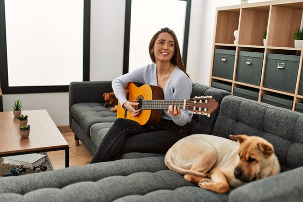 Fiatal spanyol nő játszik klasszikus gitár ül kanapén kutyákkal otthon - Fotó, kép