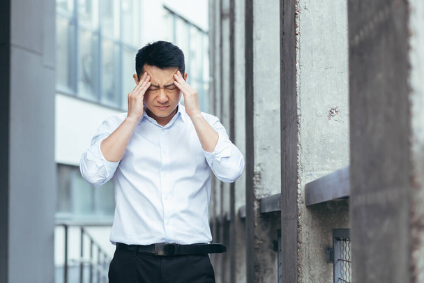Tired and sad Asian has a severe headache, a businessman outside the office, holding hands behind his head - Fotoğraf, Görsel
