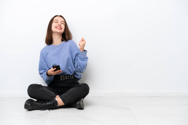 Young Ukrainian woman sitting on the floor isolated on white background with phone in victory position - Zdjęcie, obraz