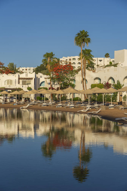 Buildings, sun loungers and parasols are reflected in the calm sea water on the beach in the resort town of Sharm El Sheikh during sunrise, Egypt - Fotografie, Obrázek