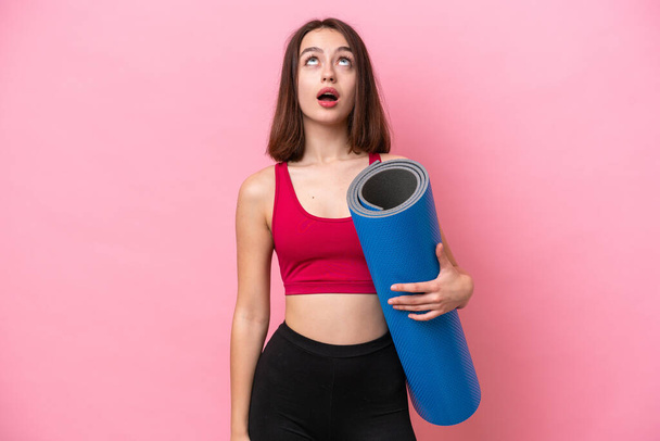 Young sport Ukrainian woman going to yoga classes while holding a mat isolated on pink background looking up and with surprised expression - Zdjęcie, obraz