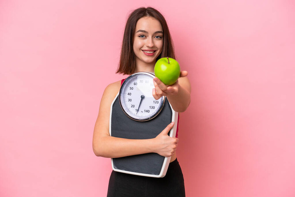 Young Ukrainian woman isolated on pink background holding weighing machine and offering an apple - Foto, immagini