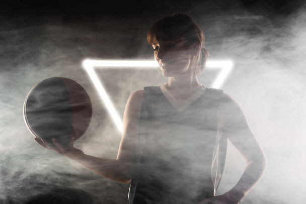 Beautiful girl with basketball. Side lit studio portrait against fog background with white neon lights. - Фото, изображение