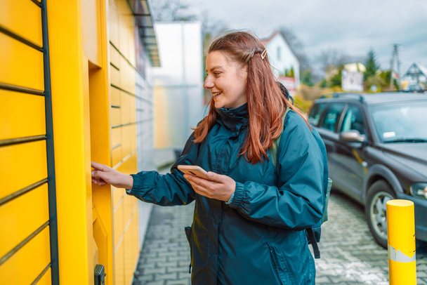 Caucasian woman showing smartphone with smart delivery app near a yellow automatic post terminal.  - Photo, Image