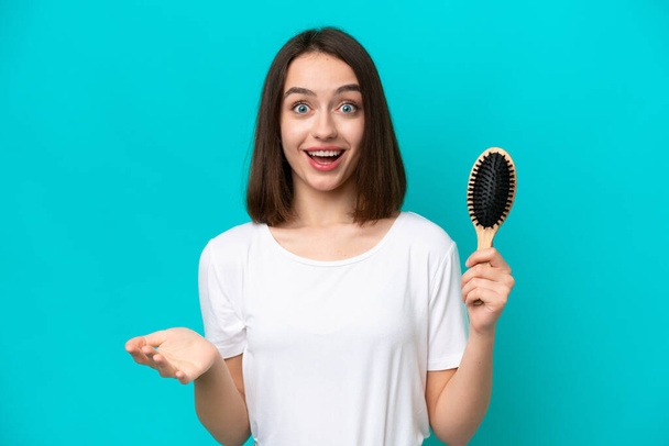 Young ukrainian woman with hair comb isolated on blue background with shocked facial expression - 写真・画像