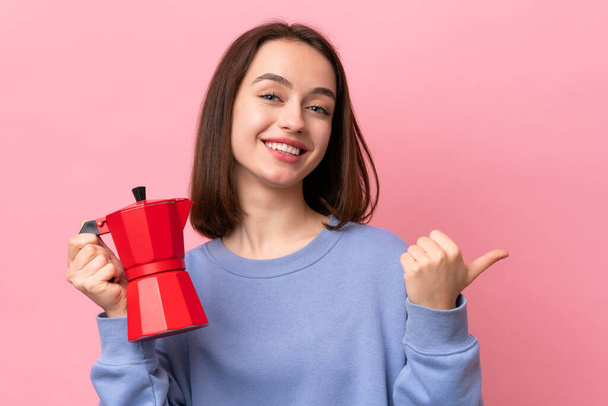 Young Ukrainian woman holding coffee pot isolated on pink background pointing to the side to present a product - 写真・画像