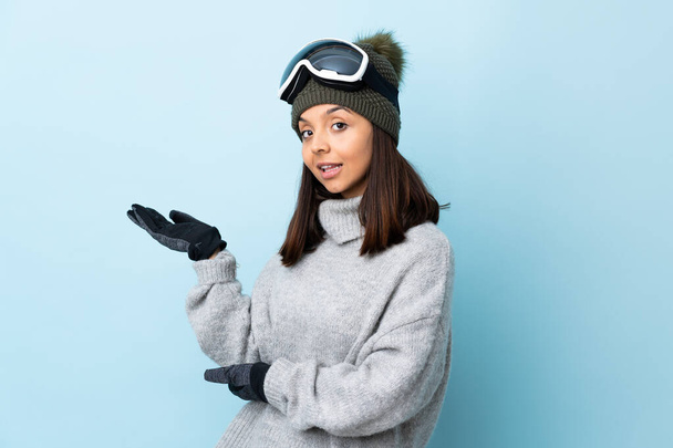 Karma kayakçı kız, snowboard gözlükleriyle izole edilmiş mavi arka planda. Davet etmek için ellerini yana uzatıyor.. - Fotoğraf, Görsel