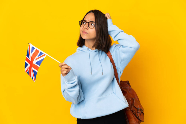 Young latin woman holding an United Kingdom flag isolated on yellow background having doubts while scratching head - Fotografie, Obrázek
