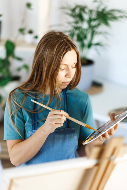 Woman artist holding paintbrush.Creative woman enjoying painting.Young beautiful woman in art studio, taking colorful paints from palette,creating masterpiece on easel, work, imagination. - Foto, Imagem