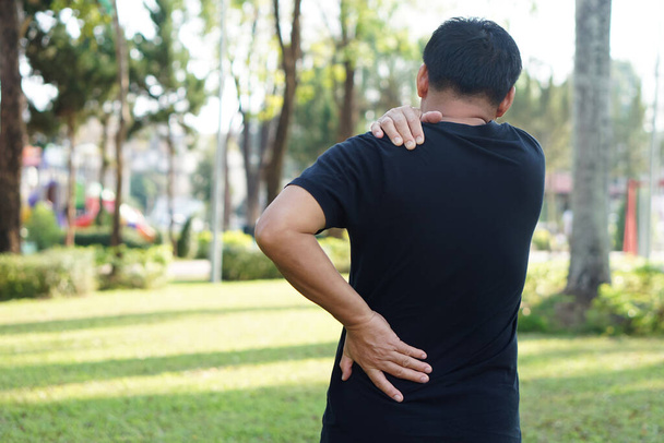 Back view of man hurts his shoulder and waist at outdoor park. Concept : Health problems, swelling and inflammation, pain. Symptoms of health injury in daily life.                 - Foto, afbeelding