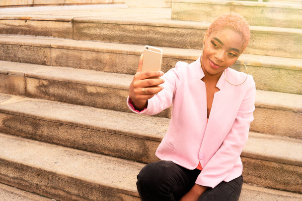 beautiful black latin woman taking a selfie photo sitting on stairs in the street  - Photo, Image