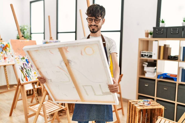 Mladý hispánský umělec muž usmívá šťastný držení plátna v ateliéru. - Fotografie, Obrázek