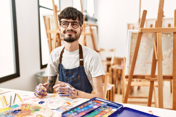 Hispanischer Mann mit Bart im Kunststudio lächelt zur Seite und starrt nachdenklich weg.  - Foto, Bild