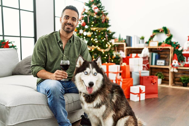 Young hispanic man drinking wine sitting on sofa with dog by christmas tree at home - Foto, afbeelding