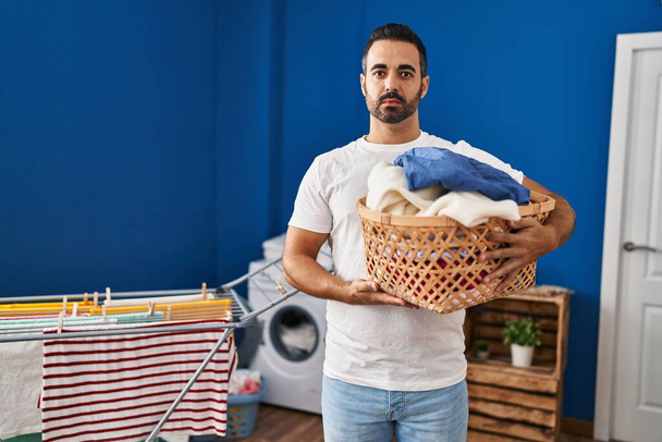 Young hispanic man with beard holding laundry basket thinking attitude and sober expression looking self confident  - Foto, afbeelding
