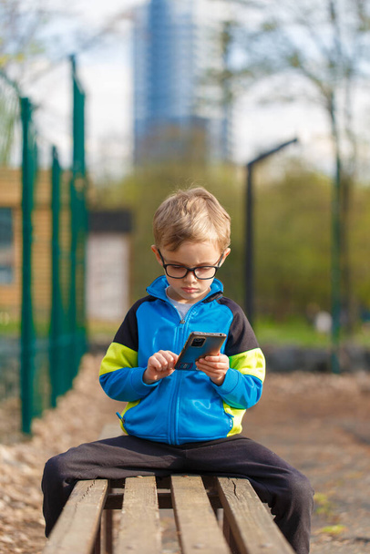 Egy ötéves, európai külsejű fiú használja a telefonot.Egy ötéves, európai külsejű fiú ül egy padon egy pvrk-ben, és használja a telefont. - Fotó, kép