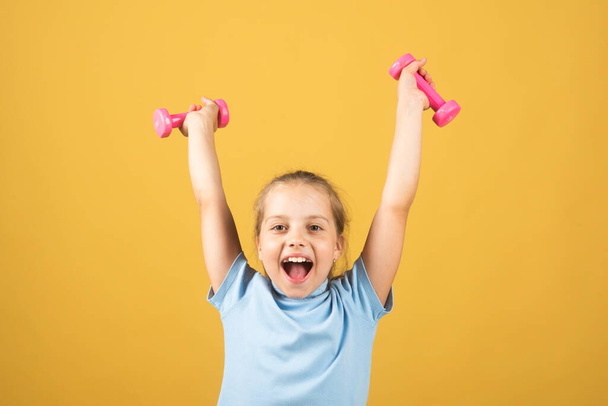 Child girl is doing exercises with dumbbells on yellow studio background. Sport portrait kids. - Foto, Imagem