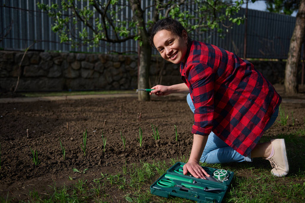 Charming middle aged Hispanic woman gardener using garden tools loosens and dogs black soil near a flower bed with growing greenery in a country vegetable garden - Valokuva, kuva