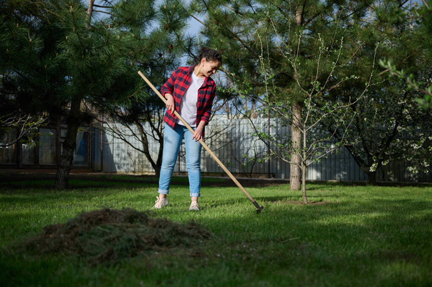 Young woman gardener cleans the local area, rakes the cut grass into a heap for further preparation of compost for fertilizing the black soil - Foto, Bild