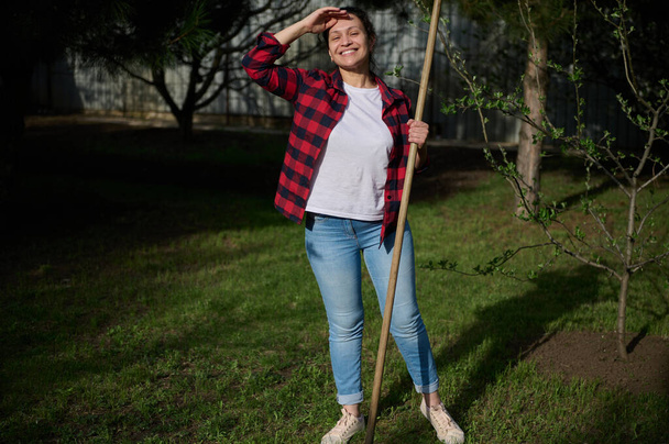 Happy female gardener standing in the backyard with a gardening tool in her hand and smiling toothy smile covering her face from the sunlight - Foto, Bild