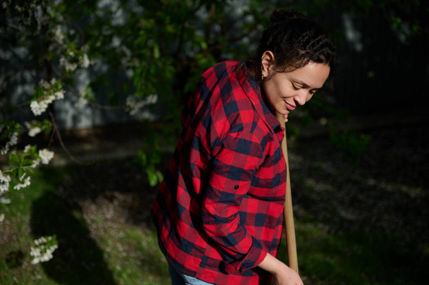 Portrait of a charming middle-aged Hispanic woman gardener housewife cleaning the local area and raking grass against blossoming fruit tree background on a sunny spring day - Photo, Image