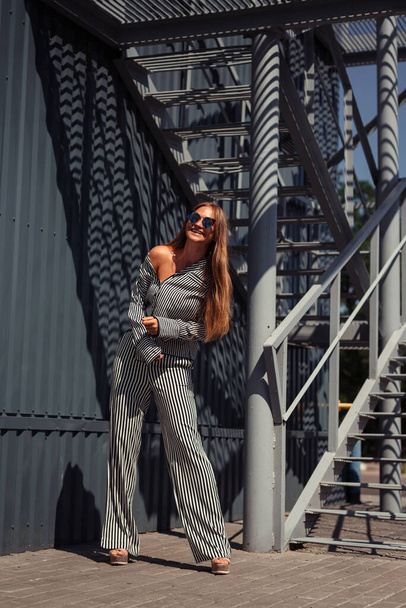 A girl in a striped suit and glasses laughs leaning forward against the background of an iron wall. - Valokuva, kuva